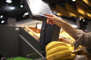 Consumidor utilizando uma balança digital em modo quiosque no supermercado para pesar frutas, com interface bloqueada para função específica.
