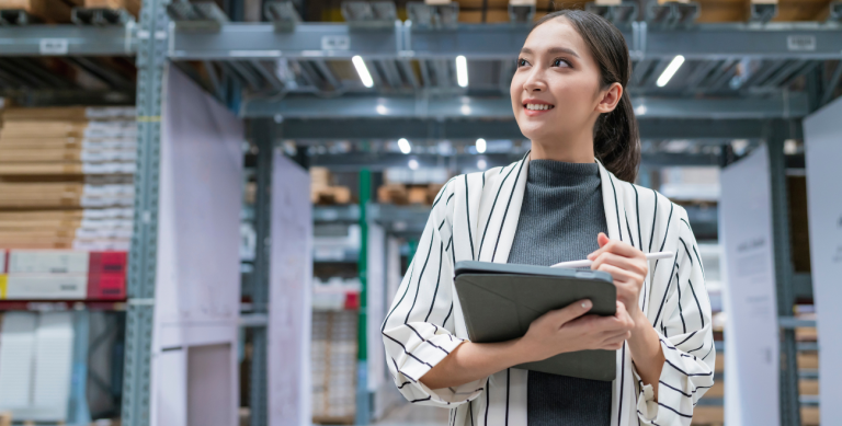 Ela olha para as prateleiras de estoque altas, representando o uso de dispositivos móveis para gestão de ativos e inventário em tempo real.